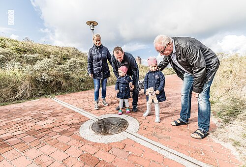 Familie auf der Juister Strandpromenade im Sonnenschein schaut sich die Markierung des 7. Längengrads an